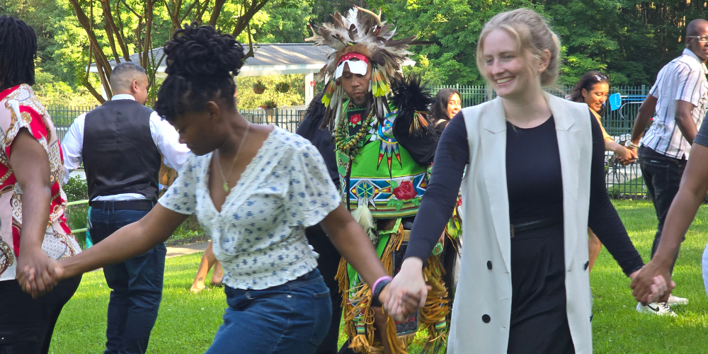 People form a circle and dance around a man in indigenous dress.