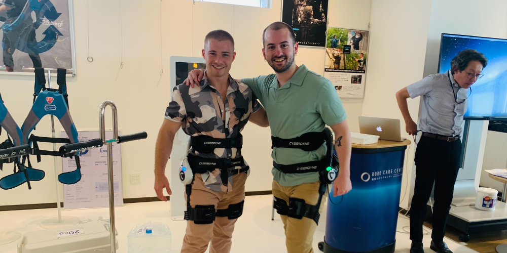 Two men wearing robotic tech pose for a photo in a Japanese lab.