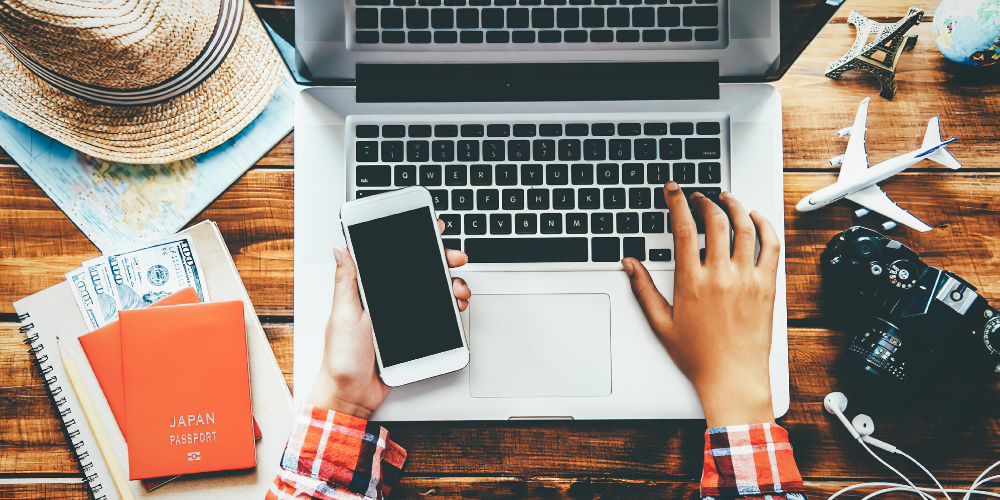 A man sits at a laptop with a cell phone in his hand and travel items on the table around him