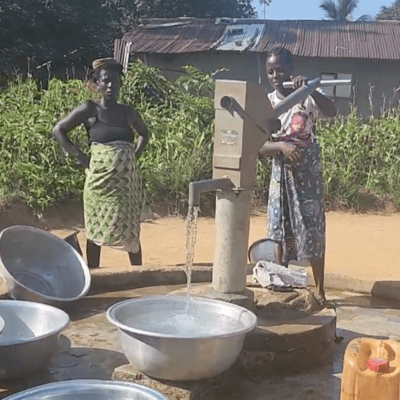 Two women use a hand pump to pump water into a large bucket