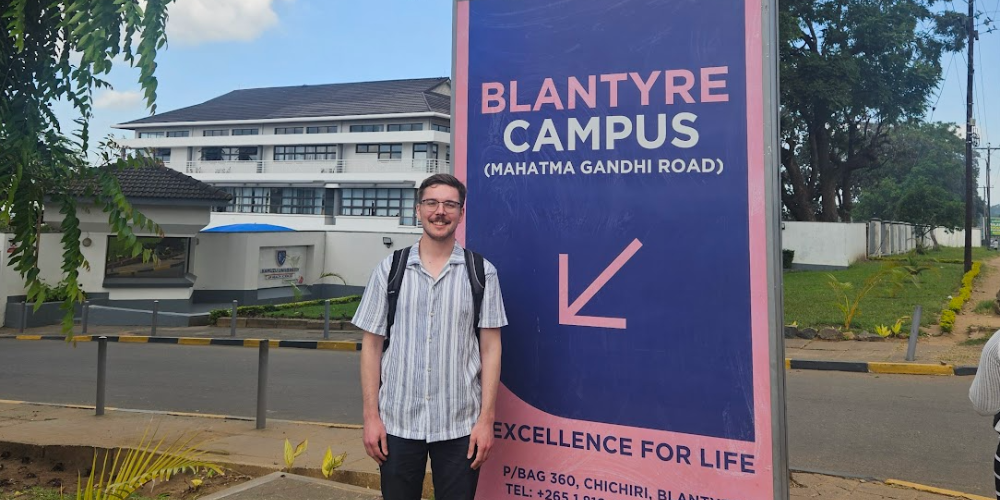 A student stands outside beside a sign for Kamuzu's Blantyre Campus