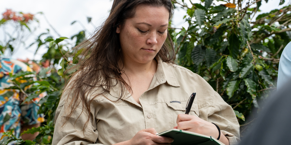 A woman writes in a journal outside with bushes behind her